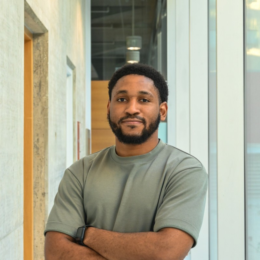 A man with a beard and short hair standing indoors near large windows with panels and natural light.