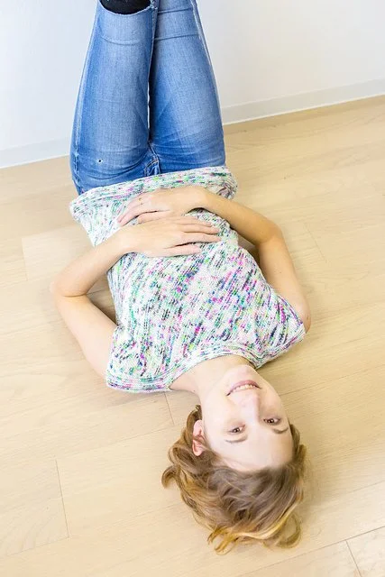 Young girl lying on her back on a wooden floor, smiling, with her head tilted sideways, wearing a colorful, short-sleeved top and blue jeans.