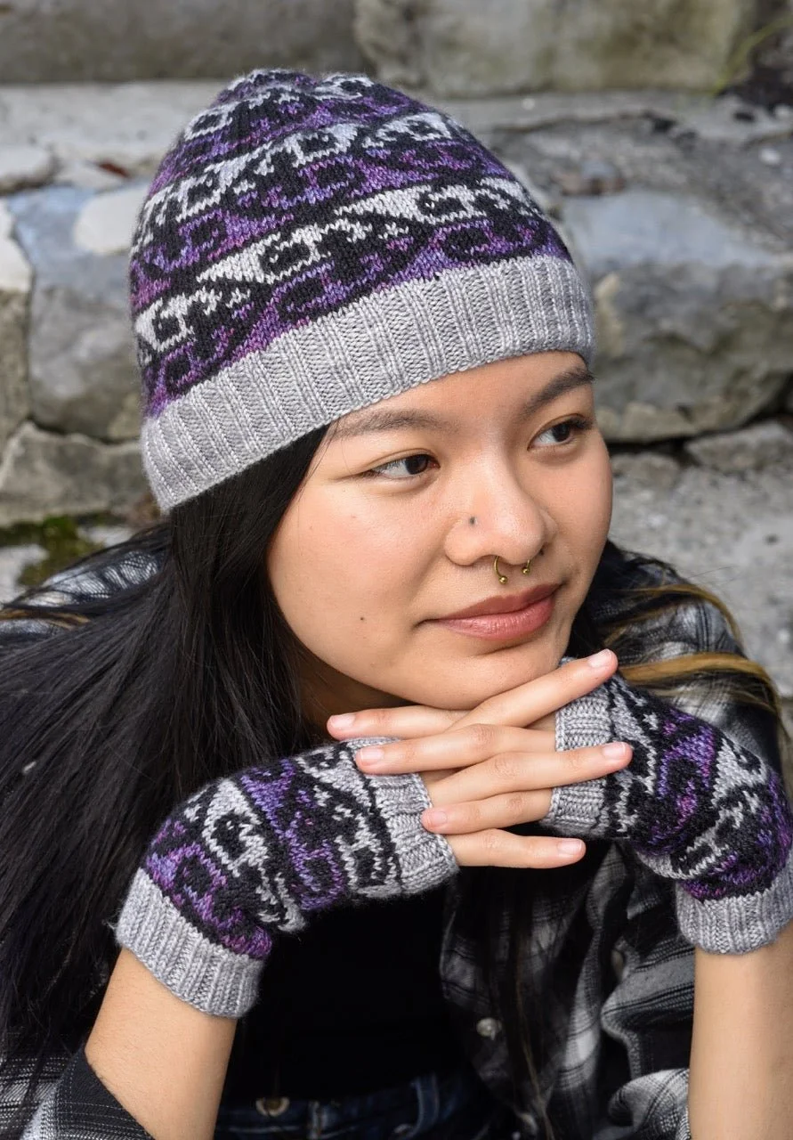 A young woman wearing a gray and purple knit beanie hat, matching fingerless gloves, and a black jacket outdoors with rocks in the background.