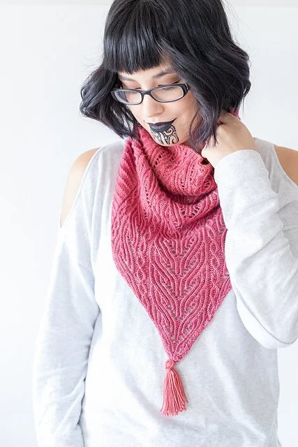 A woman with black hair, glasses, and a tattoo on her chin, wearing a gray shirt and a red knitted scarf, looking down with her eyes closed.