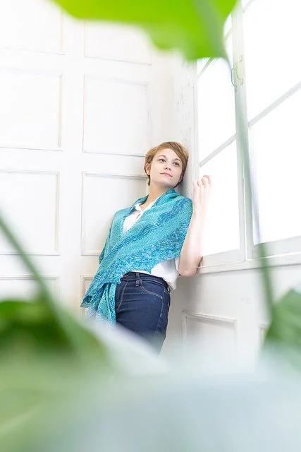 Young woman leaning against a white wall near large windows, surrounded by green plants.