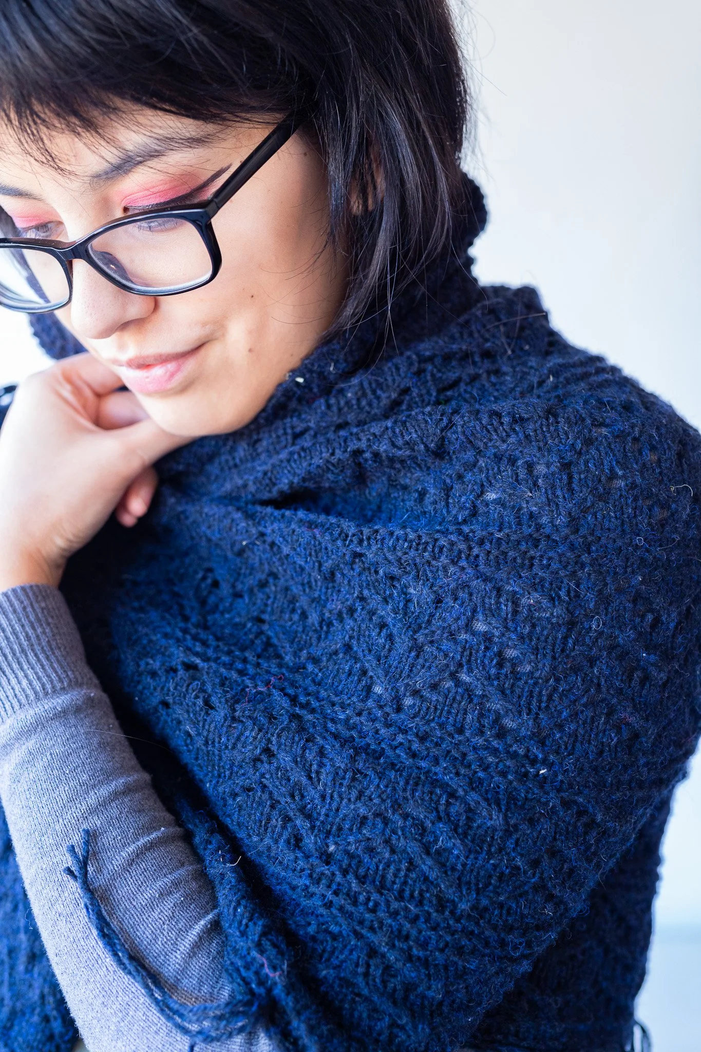 Close-up of a woman with short dark hair, wearing glasses with black frames, pink eyeshadow, and a dark blue textured sweater, looking down with a gentle smile.