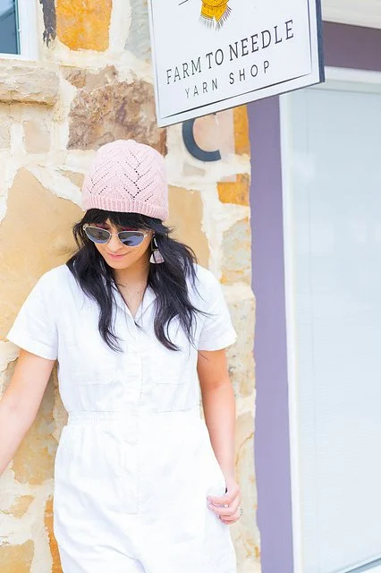 Woman in white jumpsuit, pink knit hat, and sunglasses leaning against a stone wall outside a yarn shop.