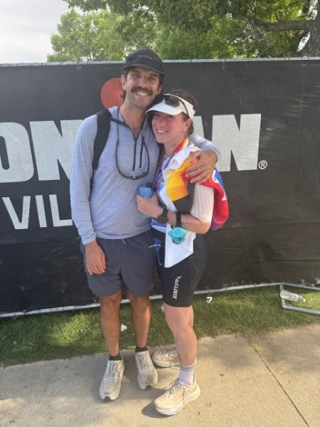 Finisher of Ironman 70.3 Boulder celebrating with her partner in front of Ironman Village sign after completing her first half Ironman.