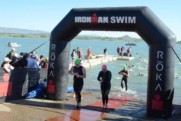 Athlete exiting the swim at Ironman 70.3 Boulder under the Ironman Swim arch after completing the 1.2 mile open water swim, with other competitiors leaving the water behind.