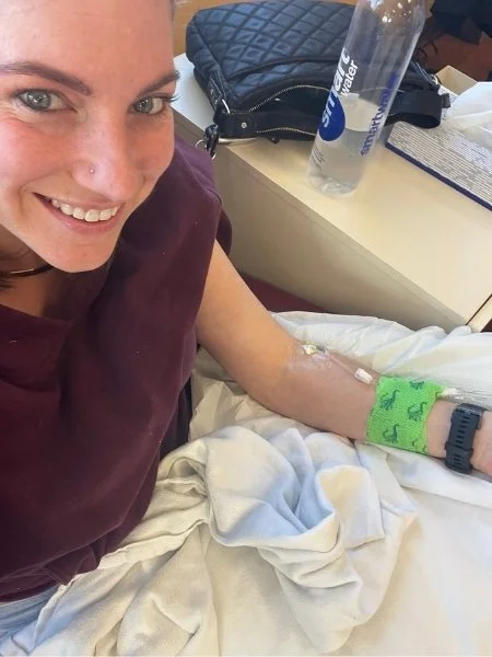 Smiling during an iron infusion while preparing for Ironman 70.3 Boulder, with IV in arm and green wrap securing the infusion site.