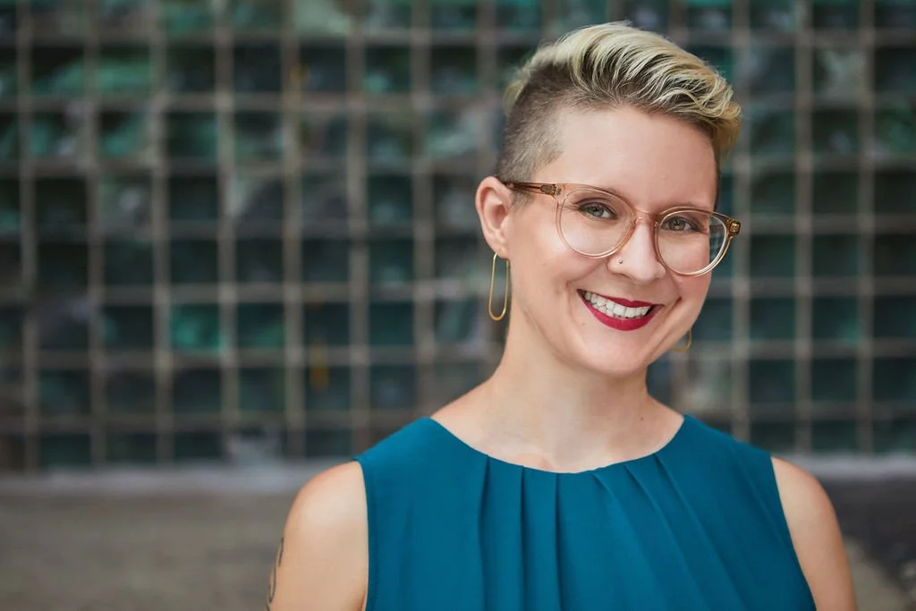 headshot of white woman in her 30s from the shoulders up, with short blonde hair and an undercut, wearing glasses and a sleeveless blue top, smiling