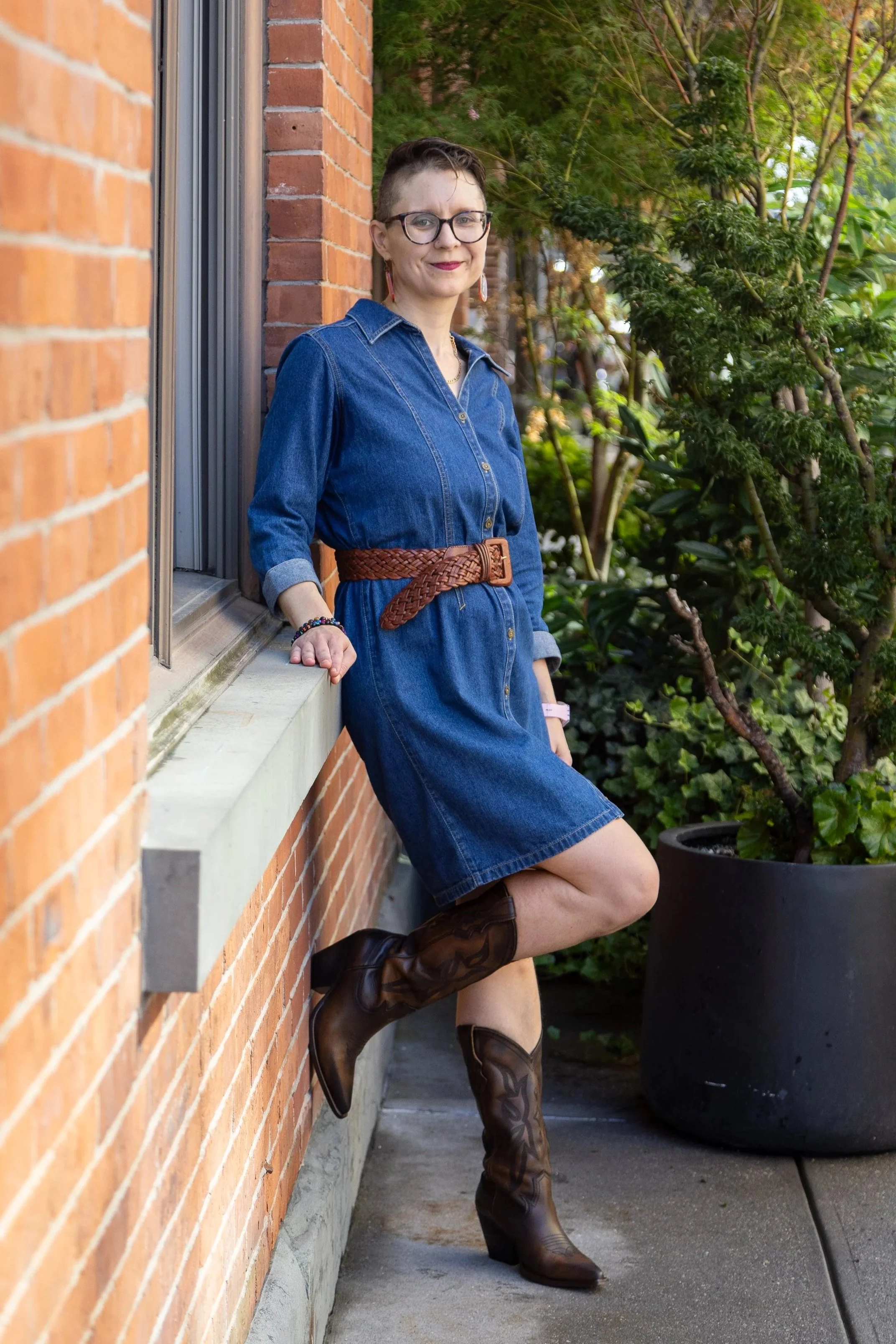 a smiling white woman, with short hair and glasses, wearing a blue denim dress and boots, is leaning against a brick wall, facing the camera