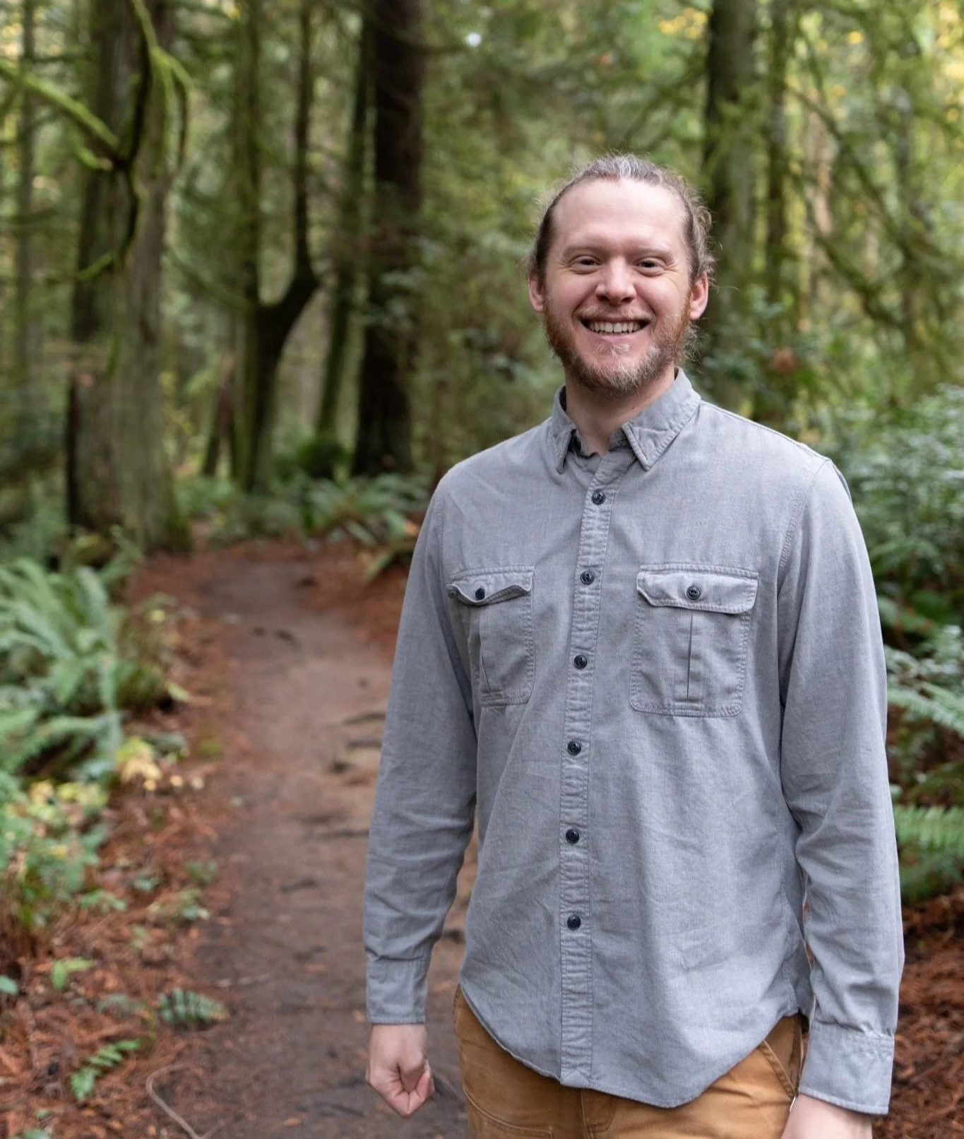 A smiling man in a gray button-up shirt stands on a forest trail surrounded by green trees and ferns.