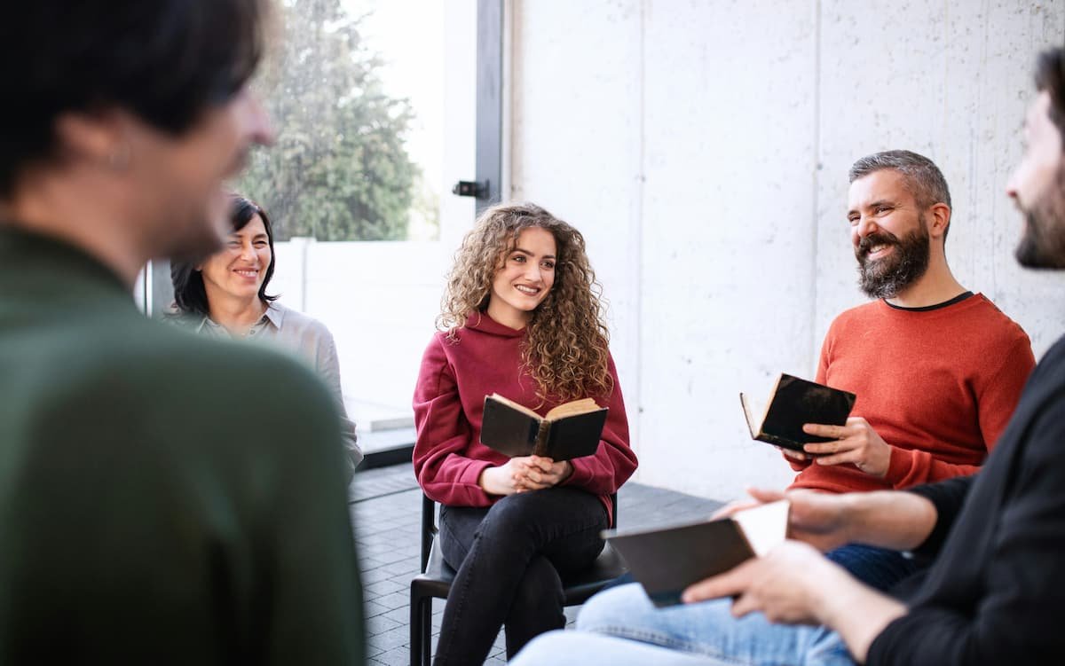 A bible study group gathered in a brightly lit room with light colored walls. A woman in a cranberry colored sweatshirt smiles as she holds a bible processing religious trauma with a man wearing an orange shirt.