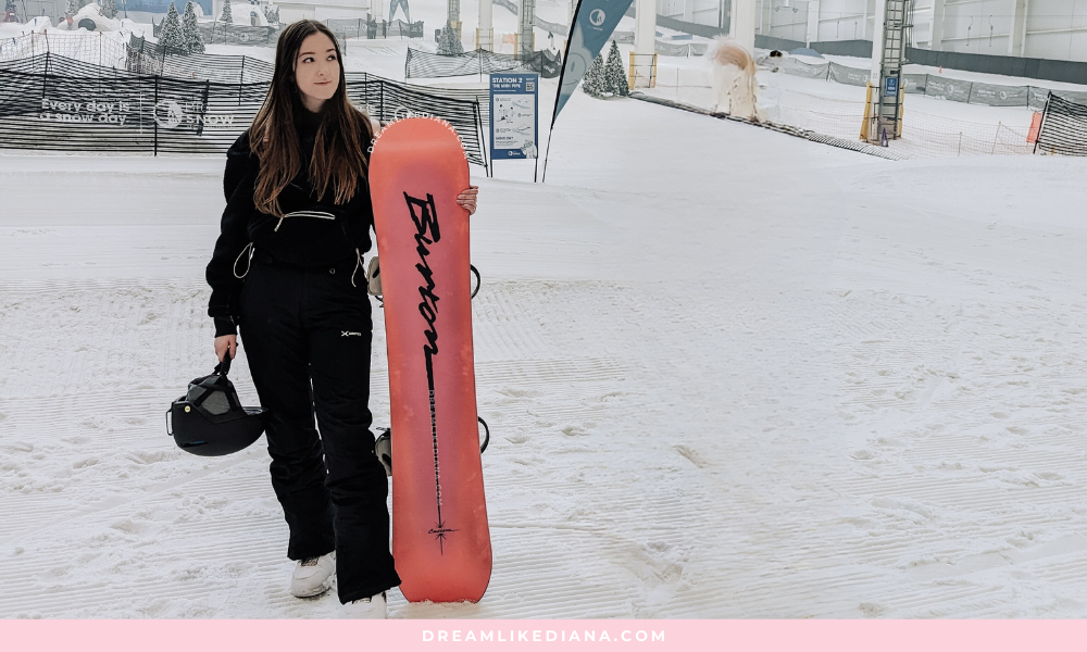 A woman in a black winter outfit stands on indoor snow, holding a pink snowboard and a helmet. The atmosphere is calm and sporty.