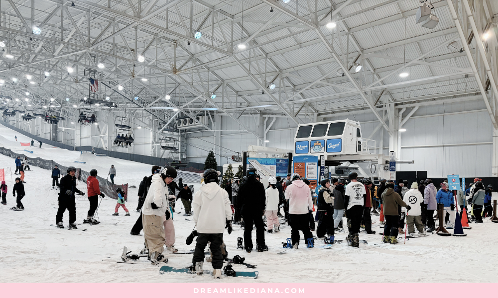 Indoor ski resort with a group of people, including children, in winter gear. Ski lift in the background. Bright, energetic atmosphere.