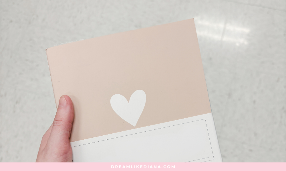 A hand holds a beige notebook with a white heart design on the cover. The background is a light, speckled floor, creating a minimalist and calm tone.