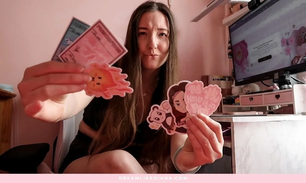 A woman in a pink room holds colorful stickers and ticket designs, featuring playful illustrations. She sits near a desk with a computer, looking playful and expressive.
