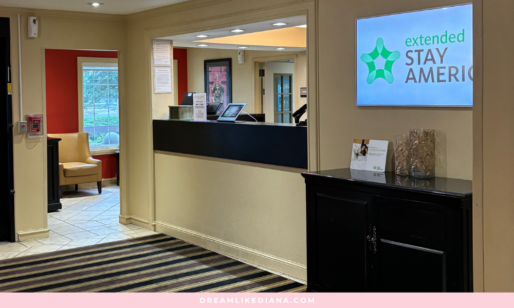 Hotel lobby with a sleek, modern design featuring a dark reception desk. Nearby is a cozy seating area with a beige chair by a window. The atmosphere is welcoming.