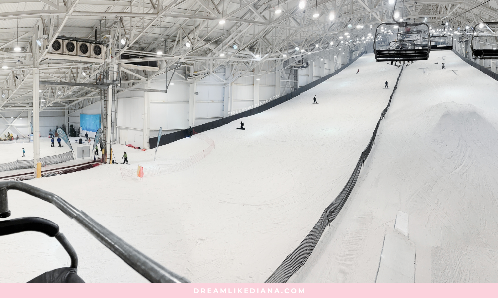 Indoor ski slope with skiers descending. Overhead lights illuminate the snowy track, surrounded by safety nets. A ski lift is visible on the right.