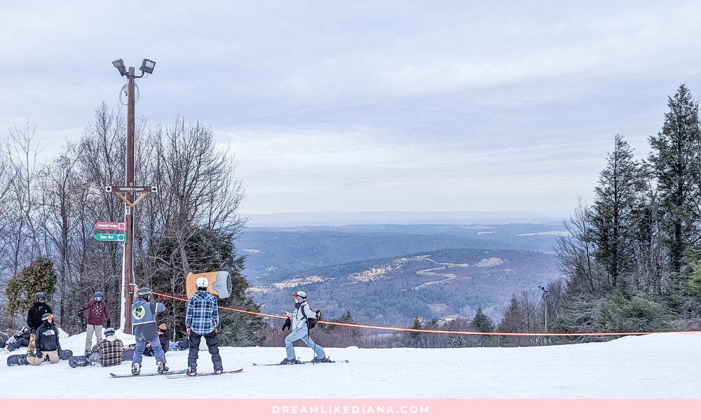 Skiers prepare to descend a snowy slope on a cloudy day, surrounded by trees. A signpost indicates trail directions. The atmosphere is tranquil.