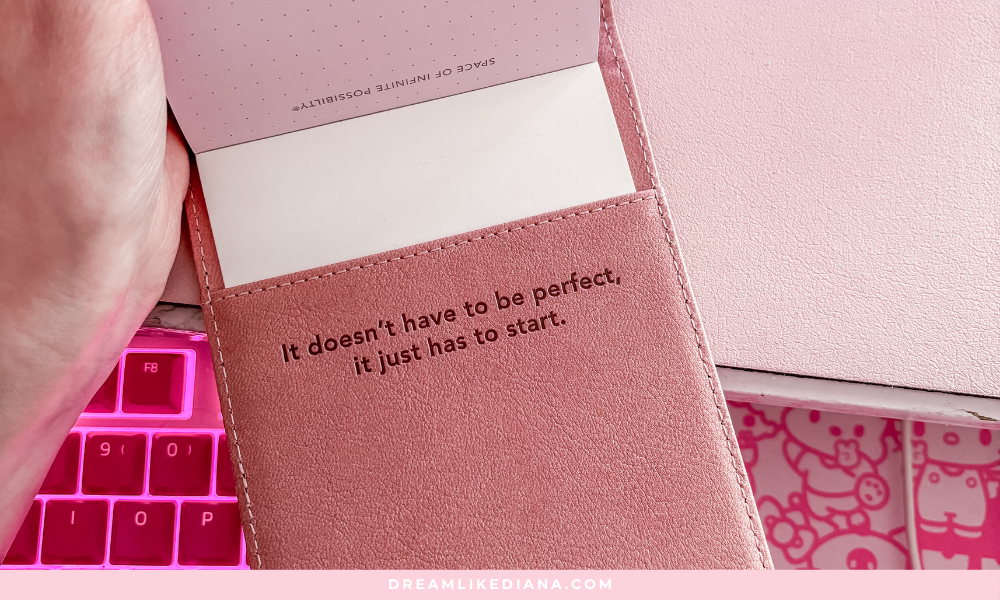 A pink notebook with the quote "It doesn't have to be perfect, it just has to start." A hand holds it above a pink keyboard and themed mousepad, conveying motivation.