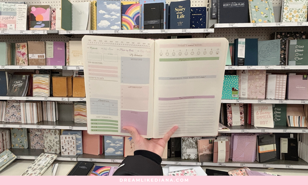 Hand holding an open planner in a store aisle filled with colorful notebooks and planners. The planner features sections for priorities, to-dos, and daily scores.