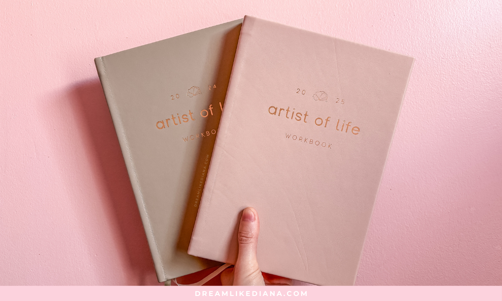 A hand holds two pastel-colored "Artist of Life" workbooks against a pink wall. The books cover 2024 and 2025, conveying a sense of calm and creativity.