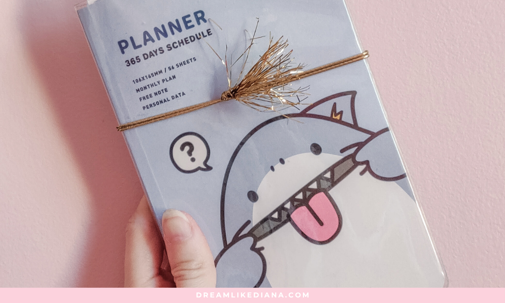 A hand holds a planner with a cute, cartoon shark illustration sticking out its tongue. The planner cover reads "Planner 365 Days Schedule." A small decorative sprig is tied to it. The overall tone is playful and whimsical.