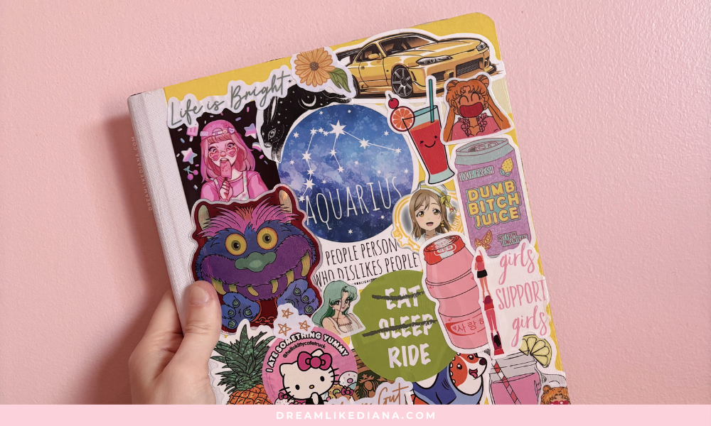 A hand holds a journal covered with colorful stickers against a pink wall. Stickers include an Aquarius theme, cars, anime characters, and motivational quotes.