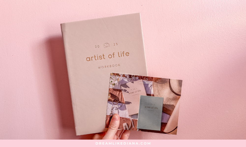 Beige "Artist of Life" workbook and colorful postcard against a pink wall, evoking creativity and inspiration. Hand holding the postcard delicately.