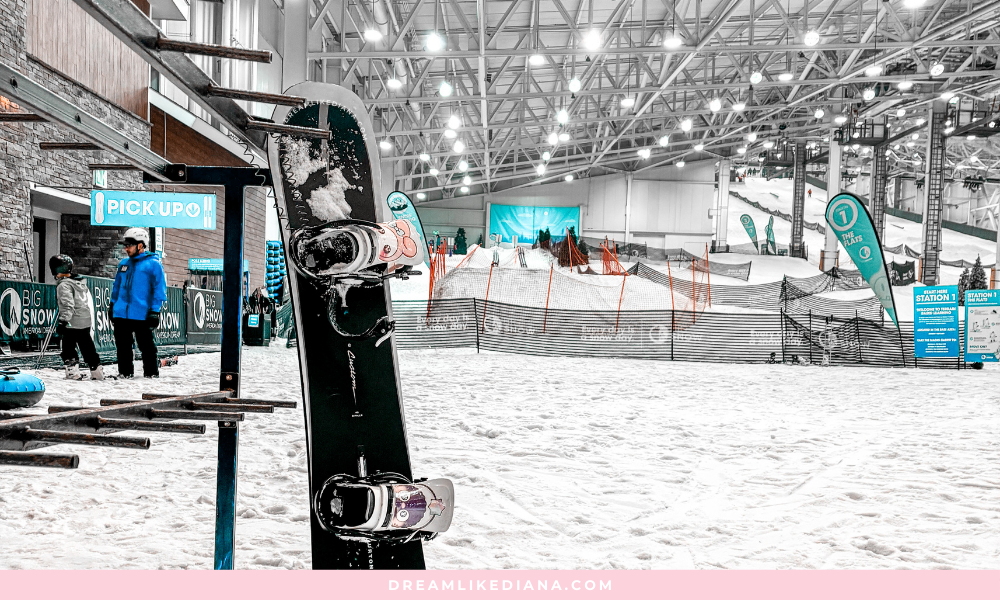 Indoor snowboarding area with a vertically standing snowboard in the foreground. People in winter attire are nearby. Bright lighting creates a lively atmosphere.