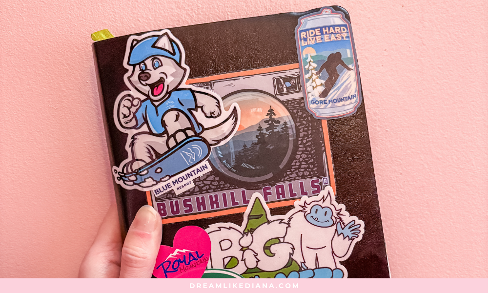 A hand holds a notebook against a pink wall, decorated with vibrant stickers: a snowboarding dog, camera lens, "Bushkill Falls," and a playful white creature.