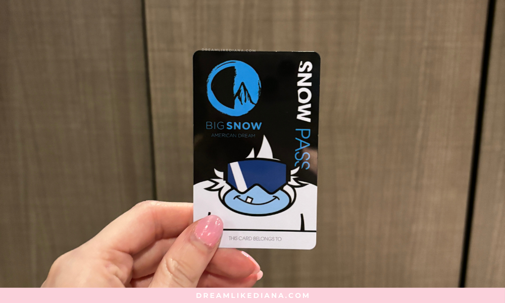 A hand with pink nail polish holds a "Snow Pass" card for Big Snow American Dream. The card features a cartoon character with goggles, set against a plain background.