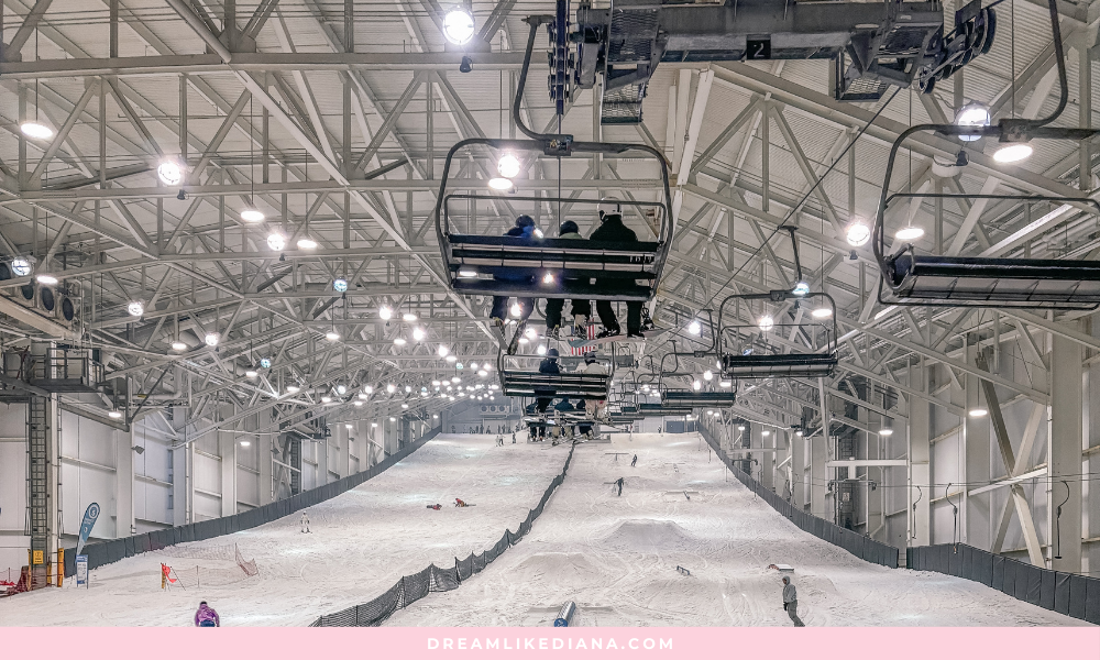 Indoor ski slope with bright lighting, showing a large space with a chairlift carrying skiers above. Snow-covered slopes, people skiing below.