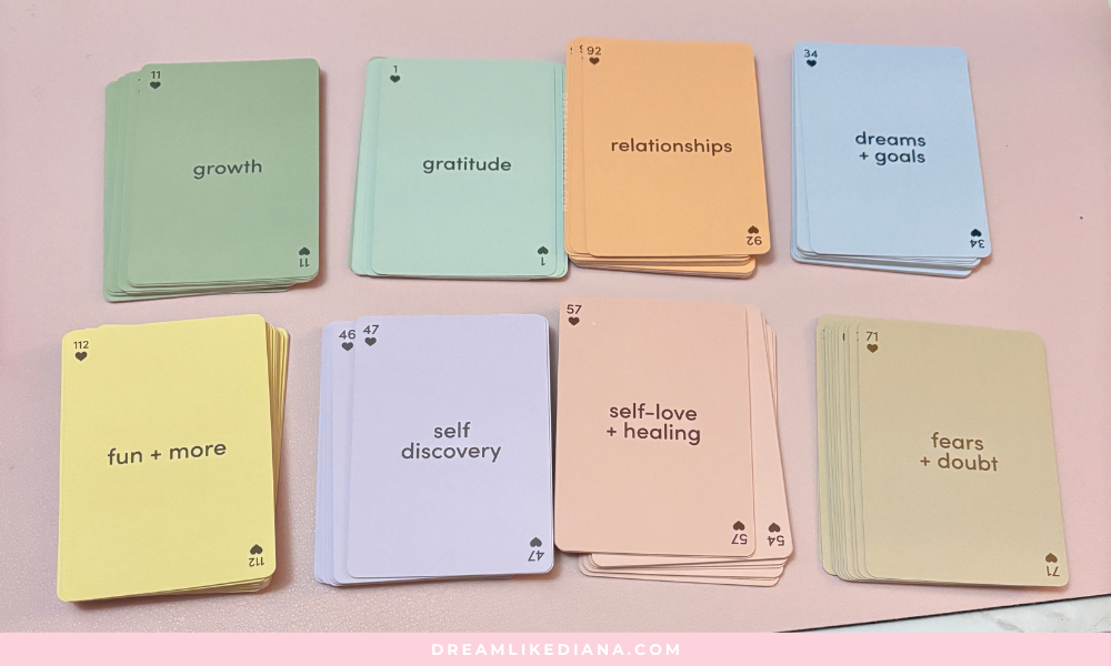 Image of pastel-colored card decks, each with different themes: growth, gratitude, relationships, dreams, fun, self-discovery, self-love, fears. Laid on a pink surface.