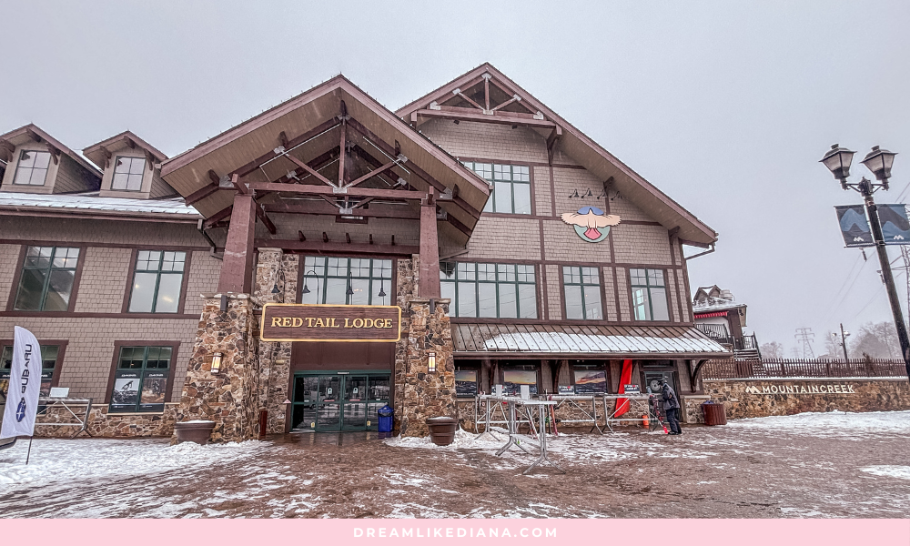 Red Tail Lodge entrance on a snowy day. Rustic stone and wood exterior, large windows, and a welcoming atmosphere. Overcast sky adds a cozy vibe.