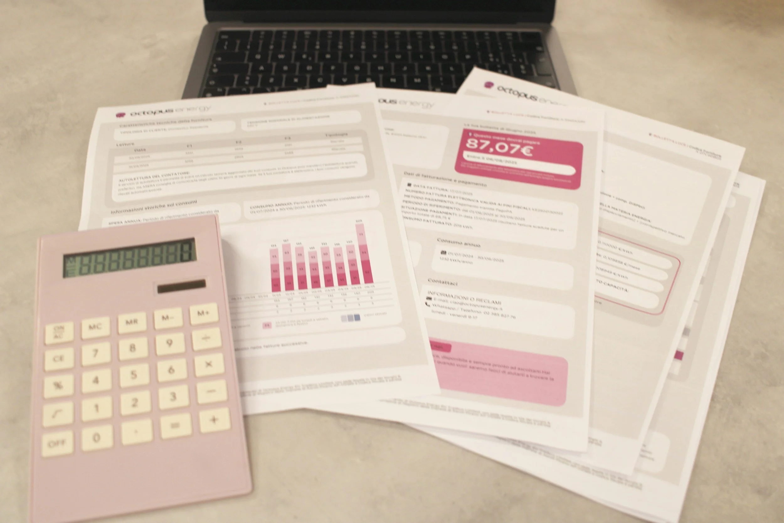 A laptop, several documents, and a pink calculator are on a desk. The documents appear to be energy bills with charts and figures.