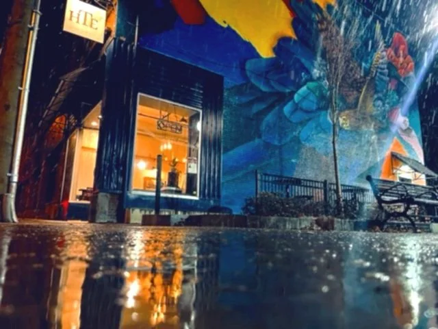 13 Central Street, Southbridge, MA - Honest Town Eats. Nighttime street scene with a mural on a building wall featuring large blue and yellow abstract flowers, a lit window with interior lighting, a sidewalk, benches, and a street sign.