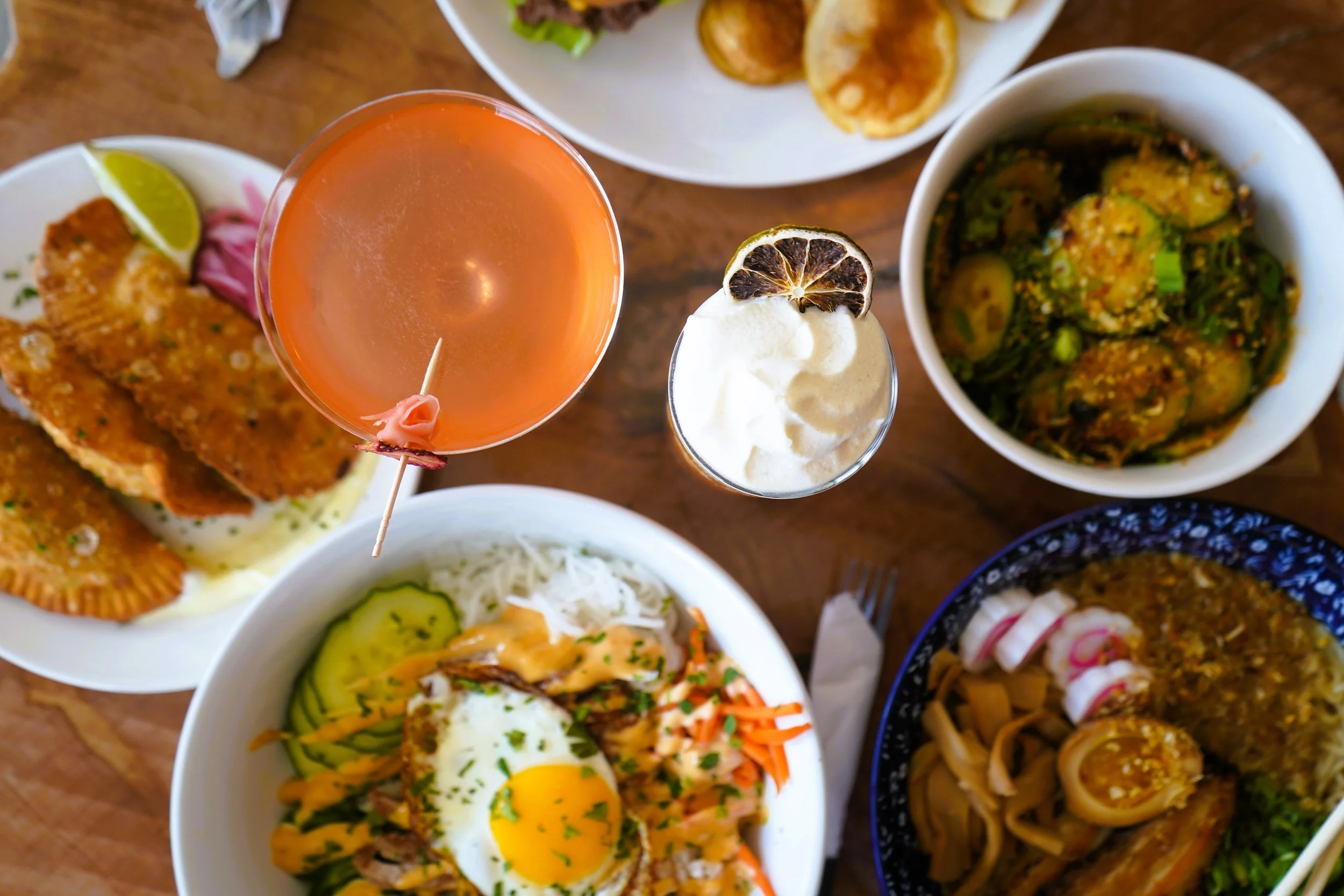 Overhead view of food from Honest Town Eats in Southbridge, MA. A table with various Asian dishes, including rice, noodles, spring rolls, a bowl of soup, a cocktail, and a dessert with whipped cream and a lemon slice.