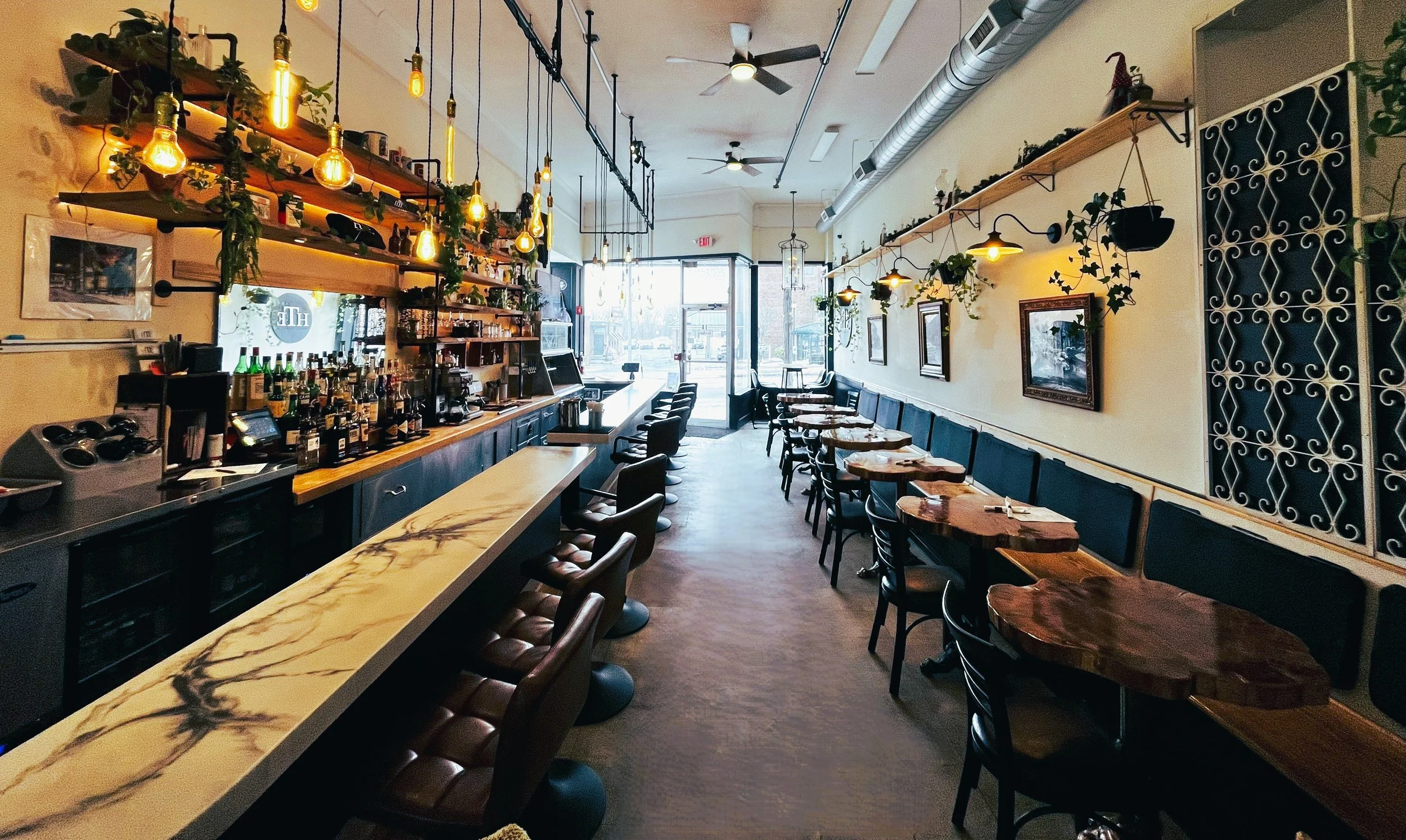Interior of Honest Town Eats, cozy restaurant with wooden tables, chairs, and ambient hanging lights, featuring a bar area on the left and a row of tables along the right wall, decorated with framed pictures and greenery.
