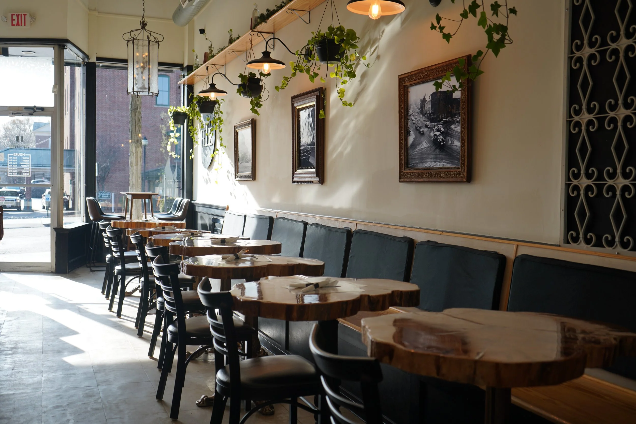 Interior of a Honest Town Eats cozy restaurant with wooden tables, black chairs, hanging decorative lights, framed black and white photographs, and hanging plants near the ceiling, illuminated by natural sunlight from the glass door and windows.
