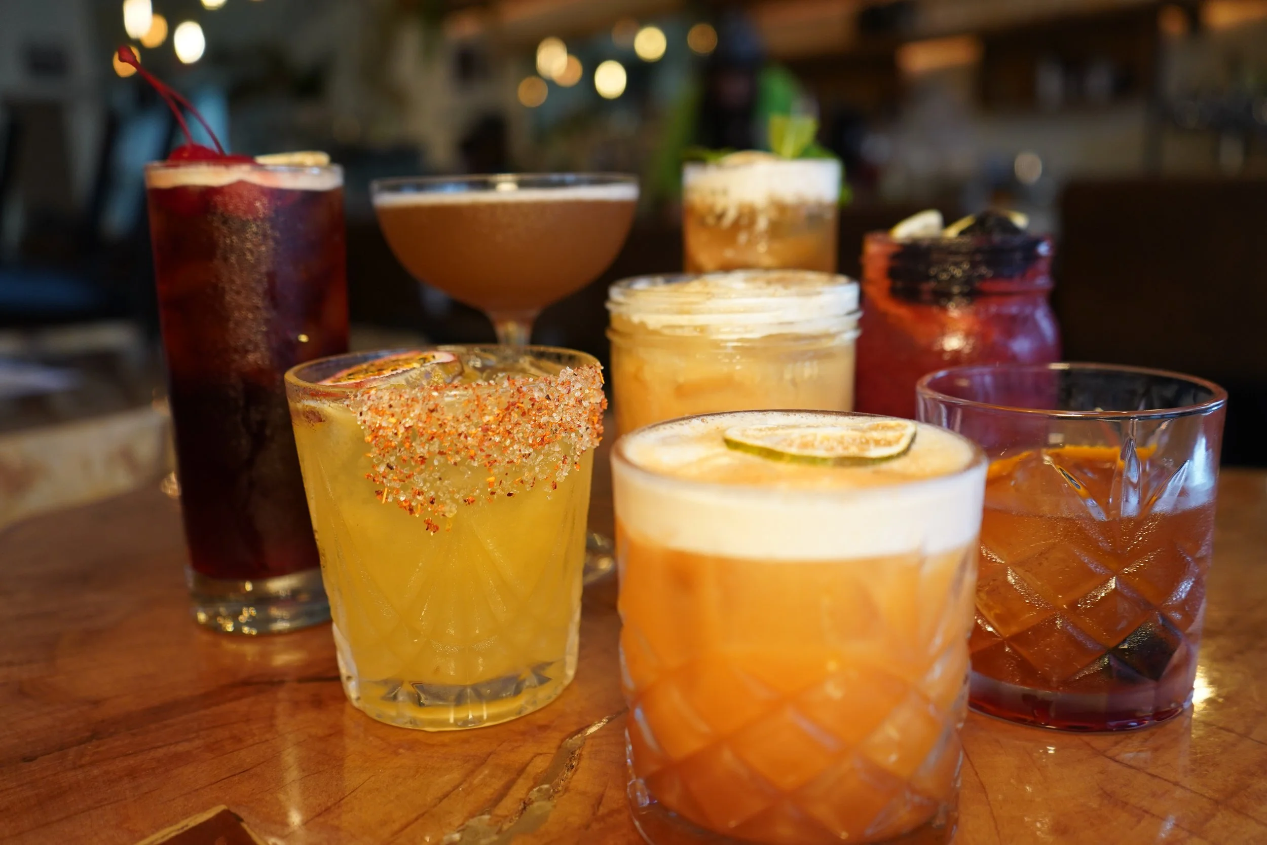 A variety of colorful cocktails and drinks from Honest Town Eats served on a wooden table in a dimly lit setting, some garnished with lime or cherries.