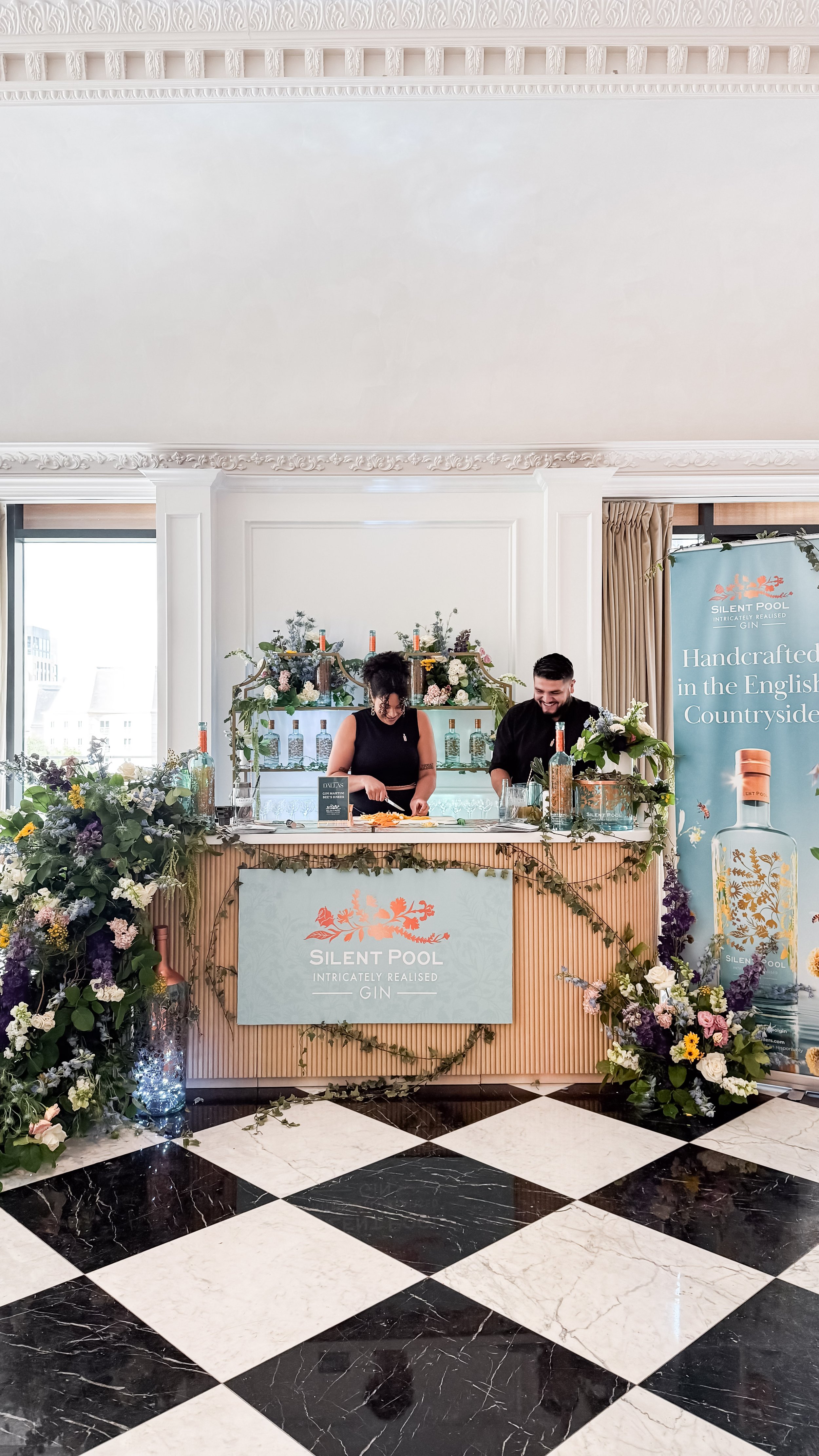 Two people preparing drinks at a Silent Pool Gin booth decorated with flowers and greenery, with a banner promoting Silent Pool Gin in the background.