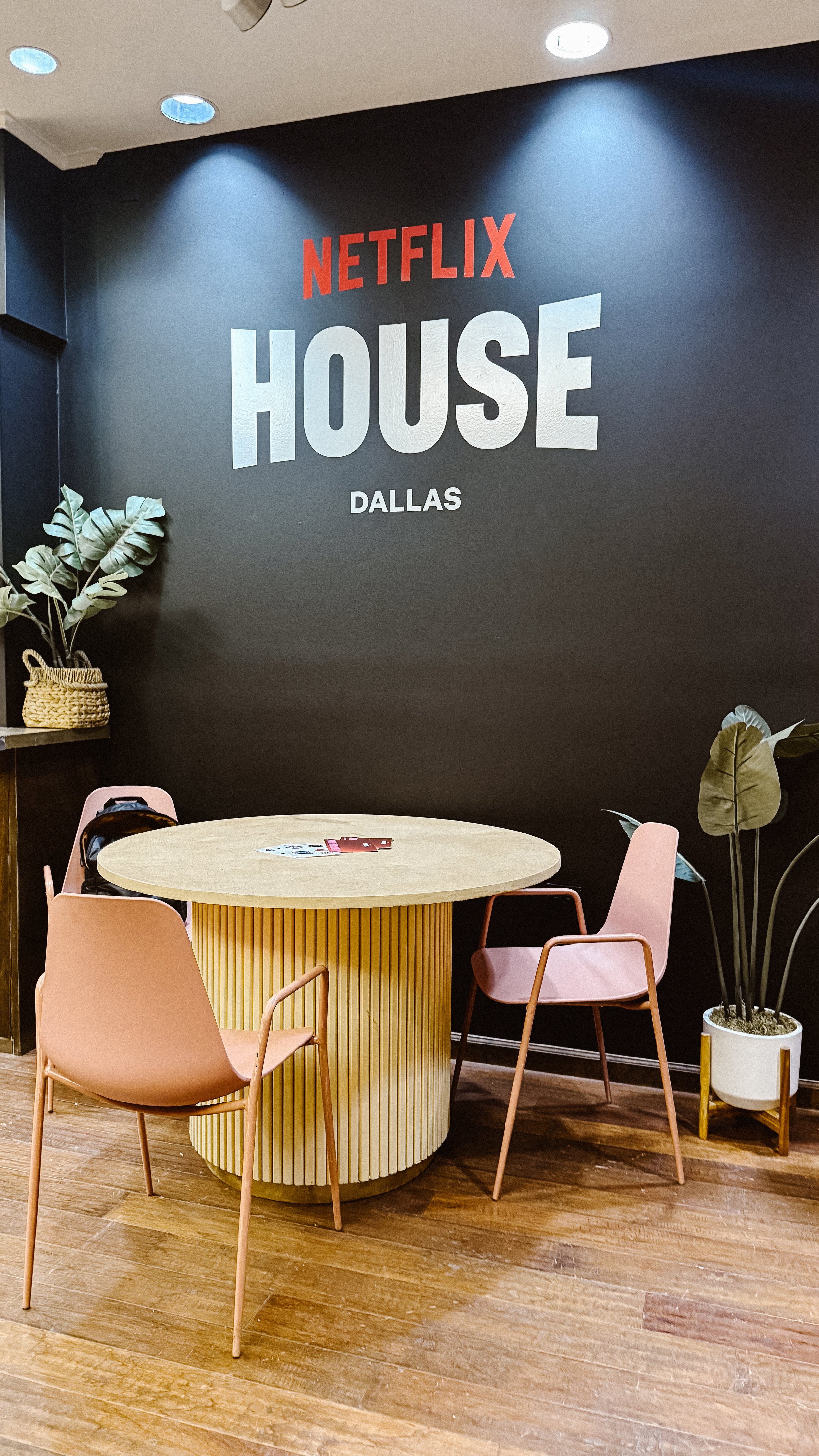 A cozy corner with a round yellow table and two pink chairs positioned around it. A black wall behind features a large sign that reads 'NETFLIX HOUSE DALLAS'. There are potted plants with broad leaves on either side of the scene, and a small stack of flyers or magazines on the table.