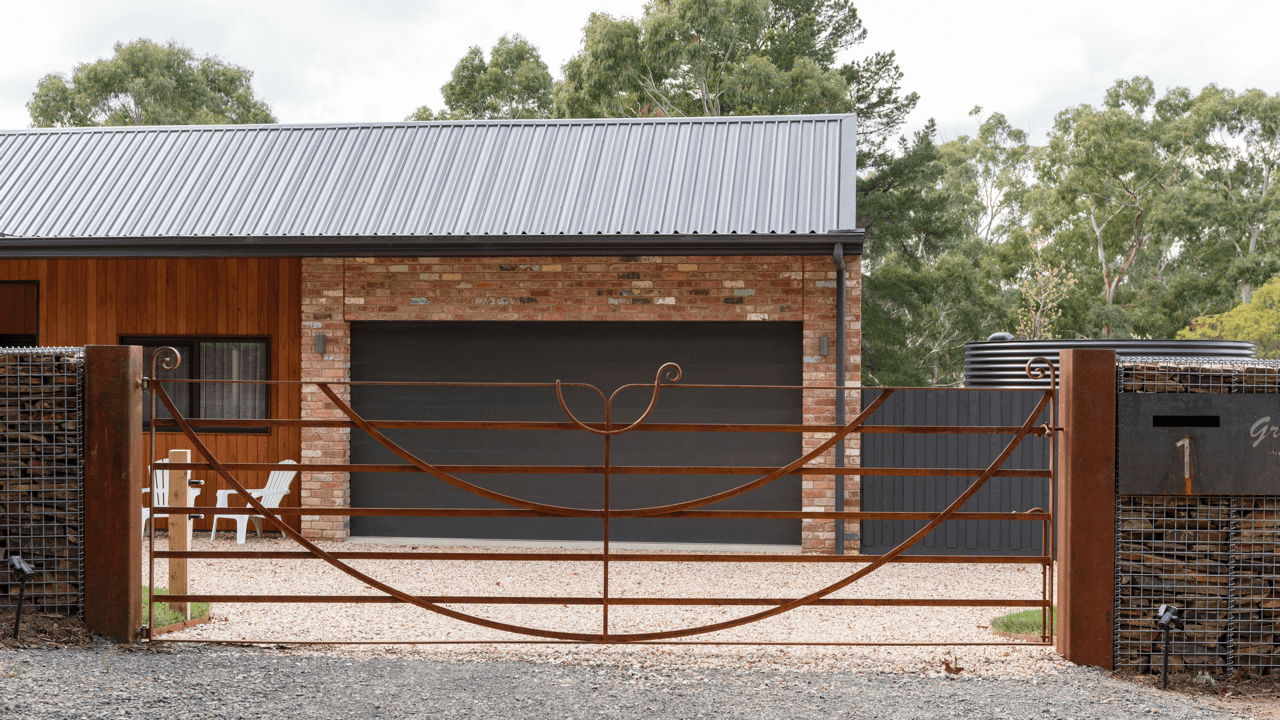 A black metal gate with weathered rusted finish and decorative curls, partially open, in front of a modern house with brick and wooden exterior, with two white chairs outside on a gravel driveway, surrounded by green trees.
