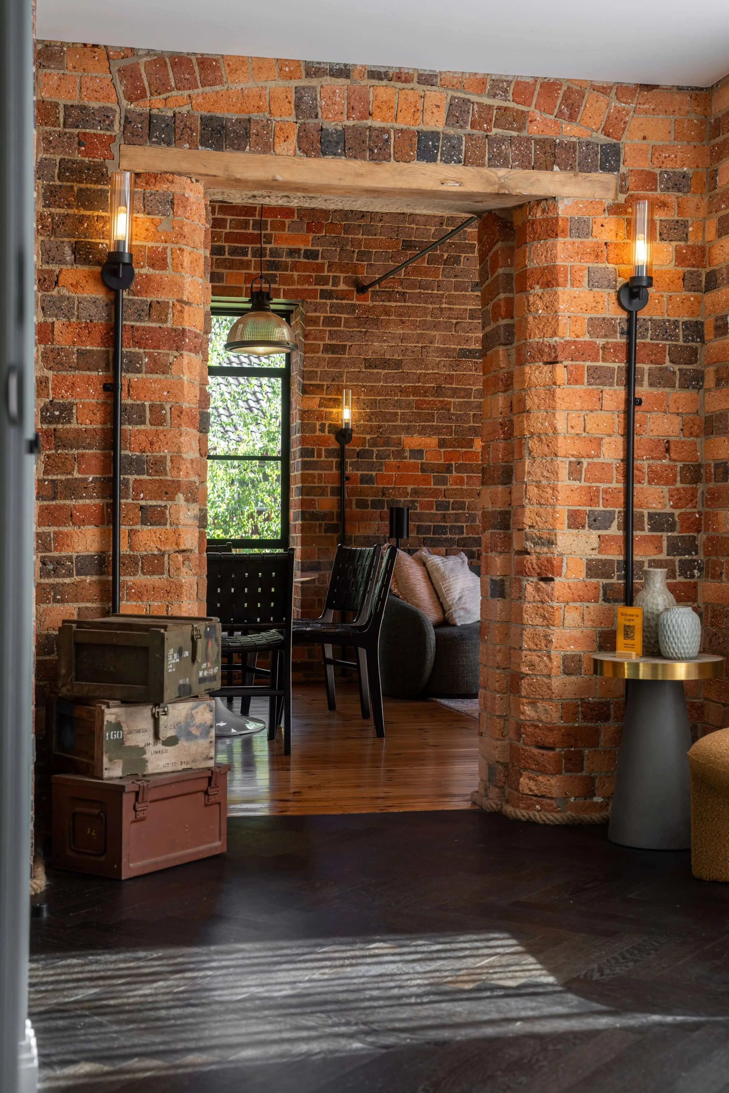 Interior of a room with exposed brick walls and wooden flooring, featuring black chairs, a sofa with pillows, hanging light fixtures, and a window with greenery outside.