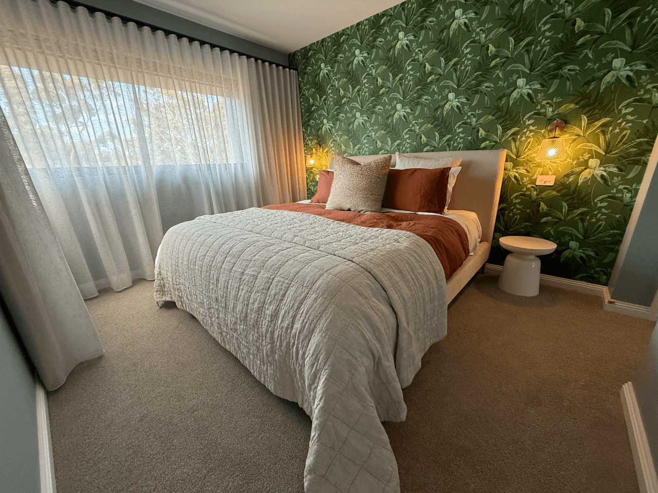 A bedroom with a bed covered in a beige quilt, pink and gray pillows, and a green floral wallpaper behind the headboard. Two wall-mounted lamps are on either side of the bed, and a small white side table is next to it. Sheer white curtains cover a la