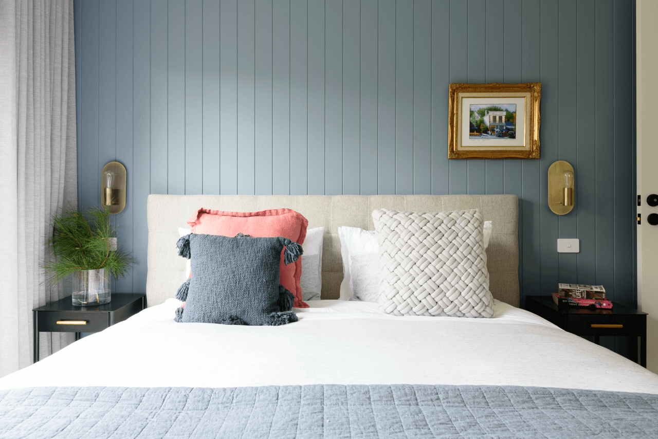 Bedroom with a white bed, decorated with three pillows in pink, dark gray with tassels, and woven white. There are black nightstands on each side, each with a book, and a potted plant on the left. The wall behind the bed is blue with a framed paintin