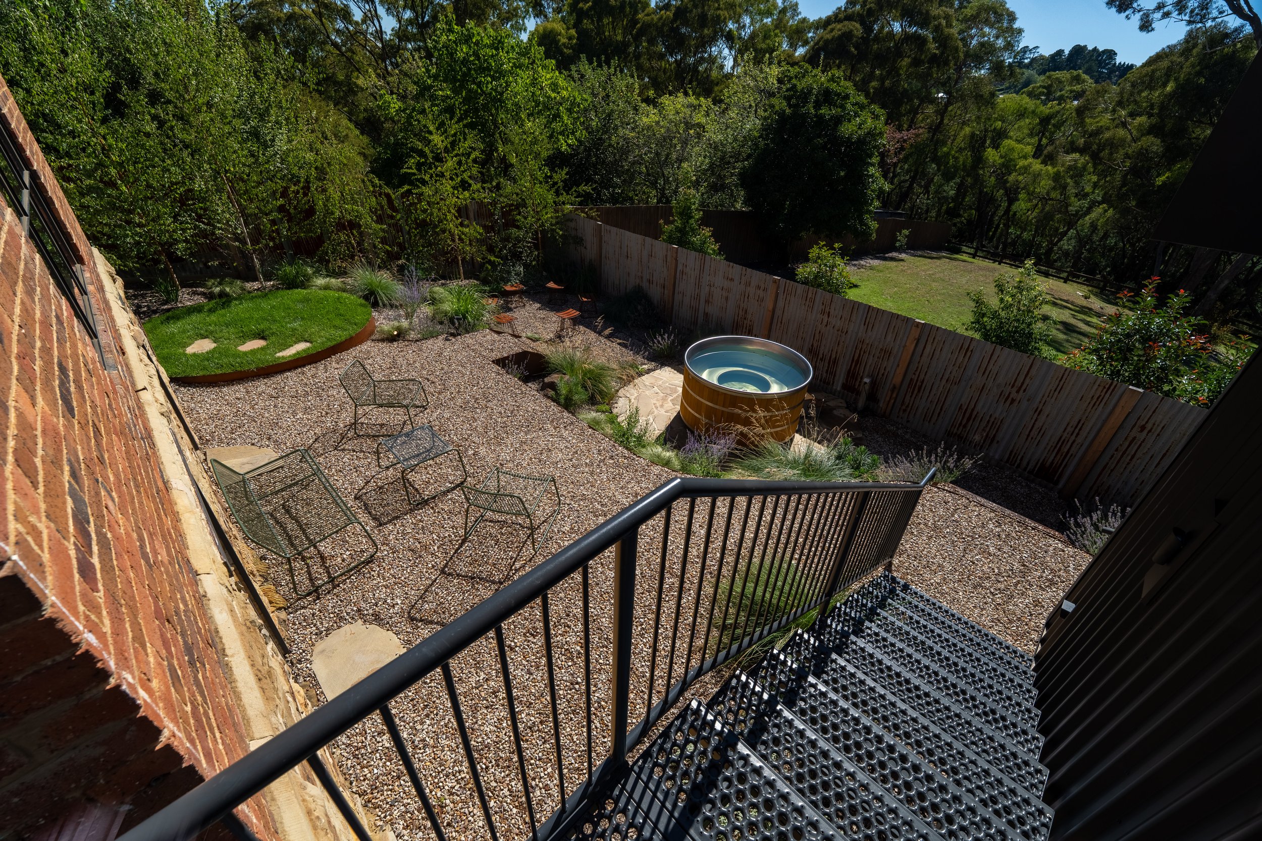 View of a backyard patio with metal chairs, a small circular hot tub, and greenery beyond a wooden fence.