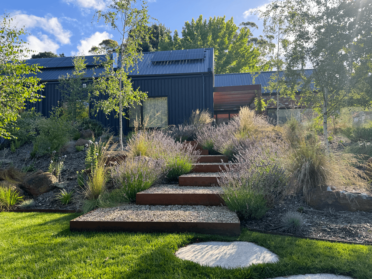 Modern house with dark blue exterior, solar panels on roof, surrounding landscaped garden with trees, shrubs, and outdoor steps leading up to the house.