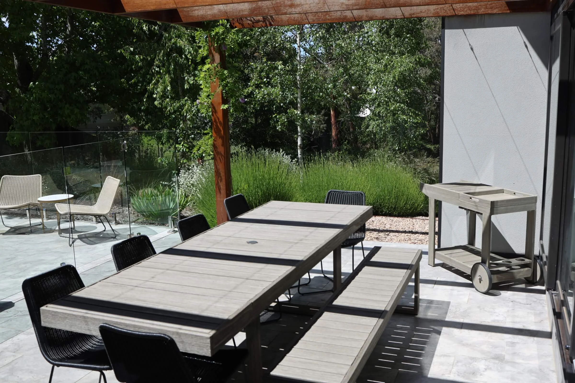 An outdoor patio area with wooden tables and black chairs, surrounded by greenery and trees, with a white building wall on the right and a sunny sky.