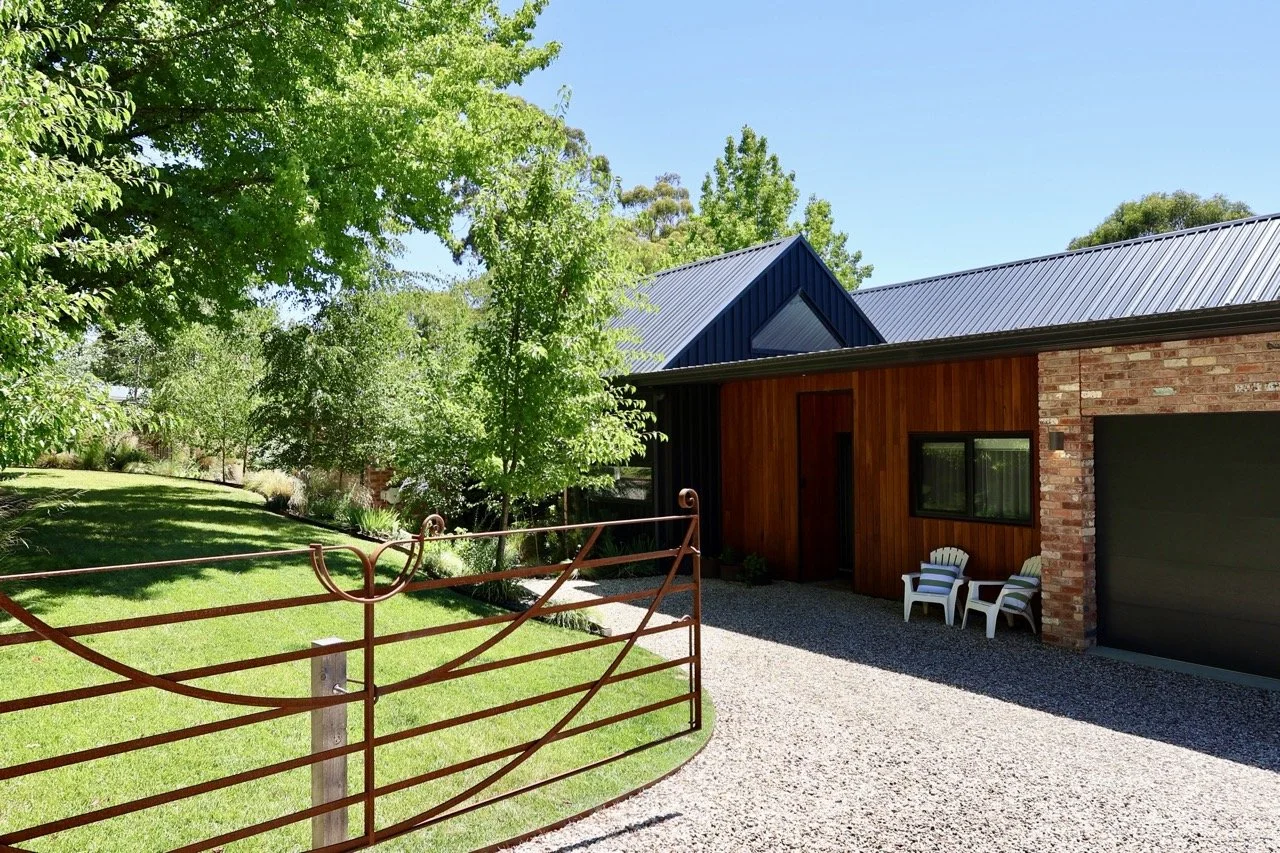 Modern house with brick and wooden exterior, green lawn, gravel driveway, and outdoor chairs, surrounded by trees and greenery on a sunny day.