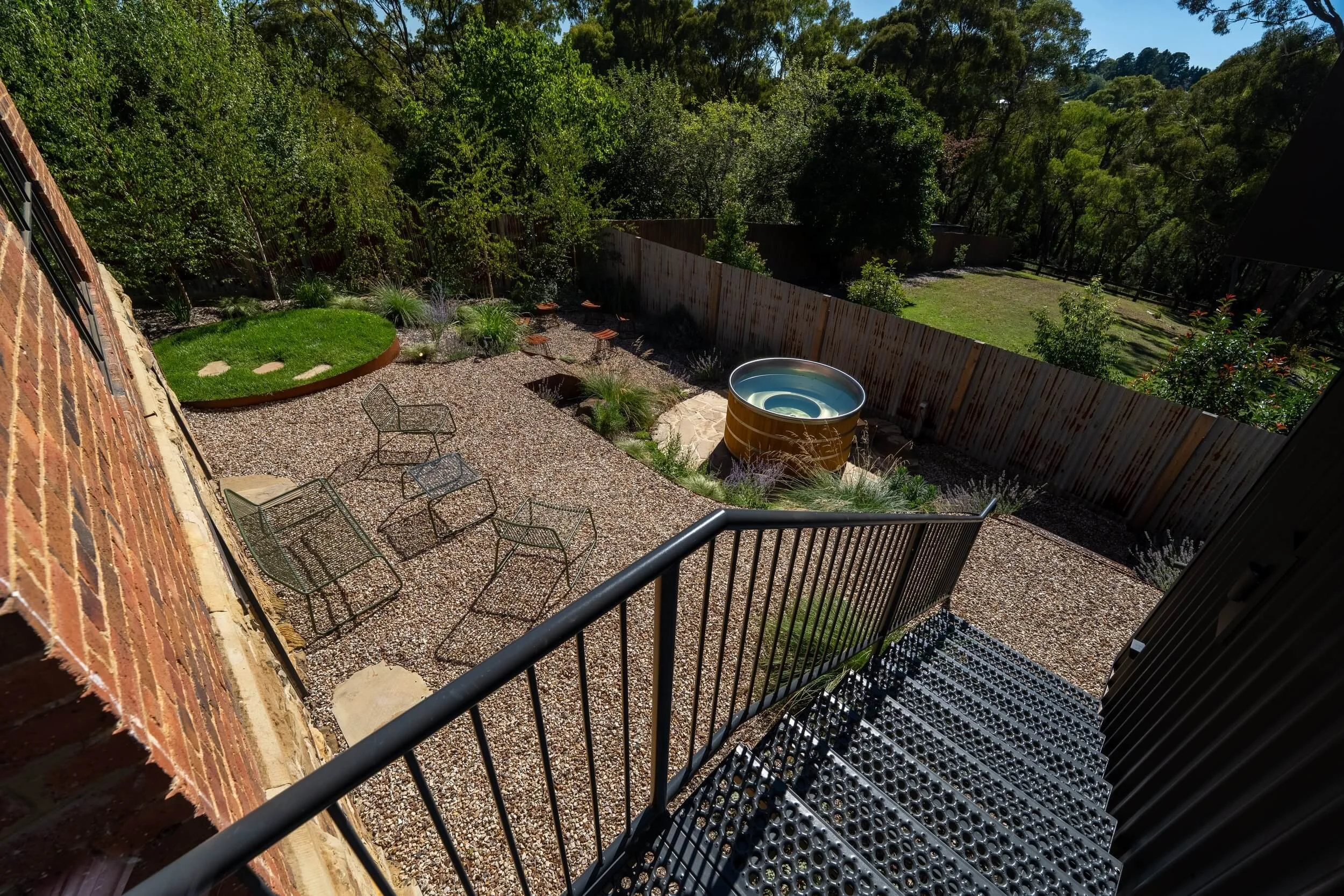 Backyard patio with gravel ground, metal chairs, a small artificial grass patch, a metal barrel pool, surrounded by a wooden fence and trees.